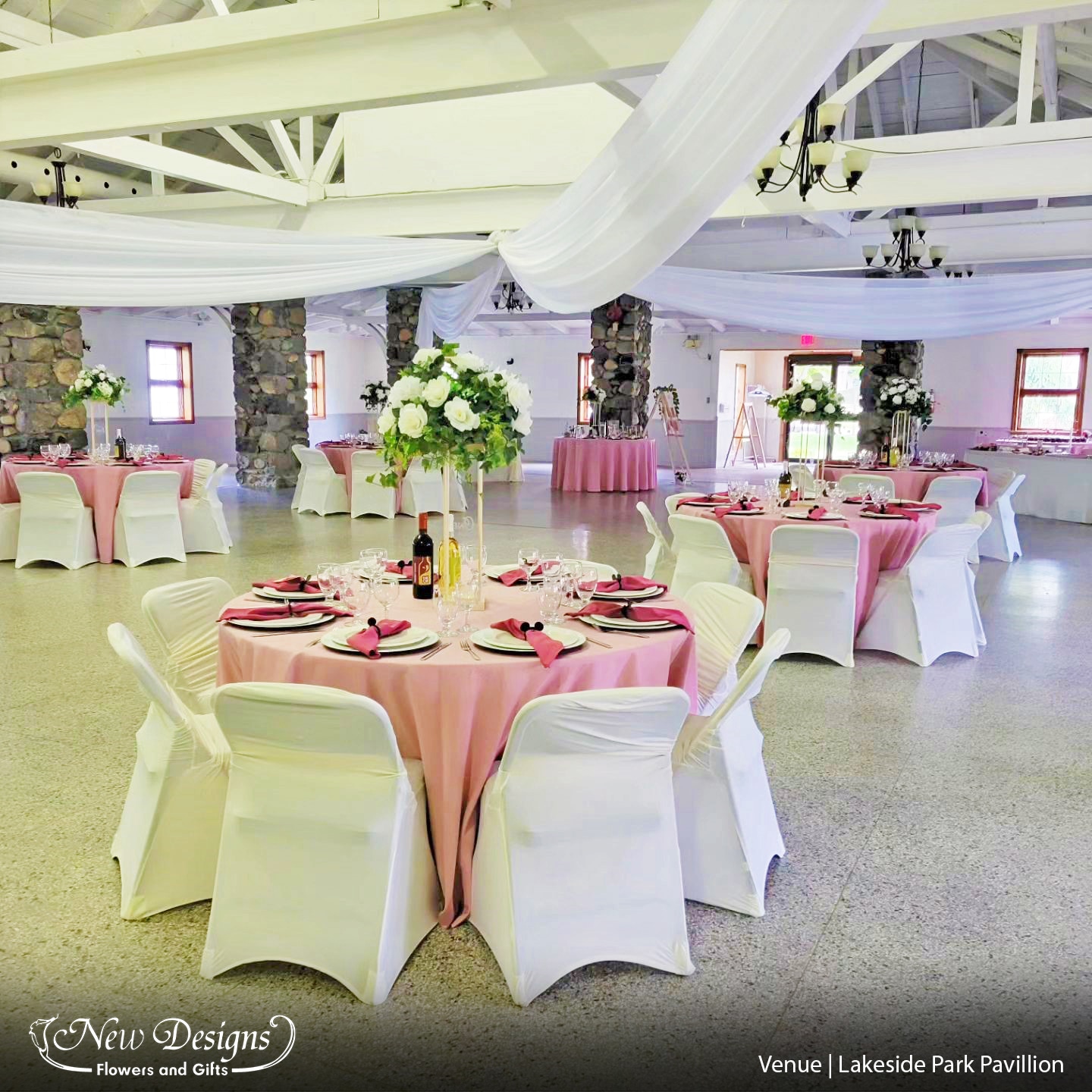 A hall in a cobblestone pavillion, White Chiffon drapes from the ceiling. Blush tablecloths line the tables, with gold charger plates, and deep pink napkins. Chairs with fitted ruffled white covers surround the table. and in the center sits a tall 4 posted wooden frame highlighted on top with a compact arrangements in whites and romantic greenery